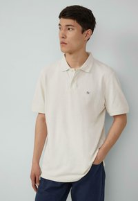 Polo shirt beige clair avec un col, des manches courtes et un logo sur le devant. Le tissu semble texturé et se porte légèrement ample sur le corps.