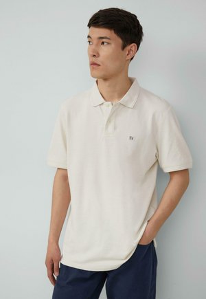 Licht beige polo shirt met een kraag, korte mouwen en een logo aan de voorkant. De stof lijkt textuur te hebben en zit losjes om het lichaam.