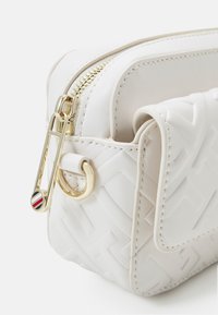 Hvid lædertaske med struktureret geometrisk mønster. Har en guldfarvet lynlås, en dekorativ guldfarvet hardware-loop og en farvet accent på lynlåsproppen.