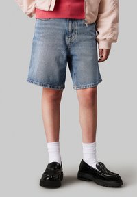 Pantalones cortos de mezclilla en un azul claro con corte recto, dobladillo enrollado y un lavado medio. Combinados con calcetines blancos y mocasines negros brillantes.