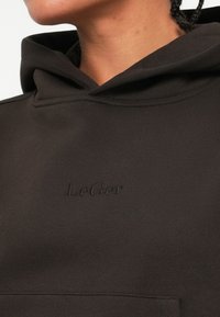 Sweatshirt preto feito de tecido macio com um bolso frontal. Apresenta um logo em relevo ao centro. O capuz tem um acabamento suave e costura mínima.