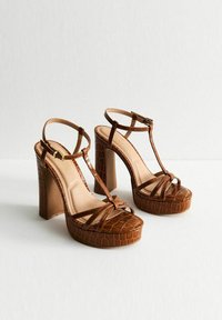 Brown faux leather platform heels with a crocodile texture, T-strap design, and chunky block heels. Features thin ankle straps and a padded insole.