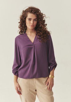 Joven con cabello rizado, vestida con una blusa morada de cuello en V y pantalones beige, posando con una pulsera dorada en la muñeca.