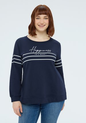 Femme souriante aux cheveux bruns mi-longs porte un sweat-shirt bleu marine à rayures blanches avec le texte « Le bonheur est une humeur », associée à un jean bleu.