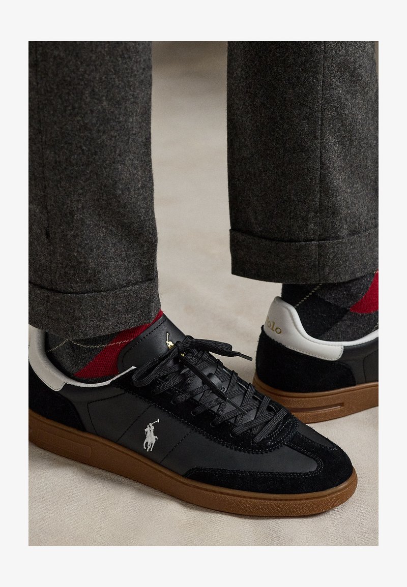 Zwarte en bruine sneakers met wit Polo-logo, gedragen met grijze en rode argyle sokken en donkergrijze omgeslagen broek op een beige vloer.