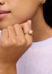 Zbliżenie na dłoń przy brodzie, noszącą złoty pierścionek z centralną perłą i złotymi perełkami jako akcentami, w połączeniu z pasującą złotą kolczykiem.