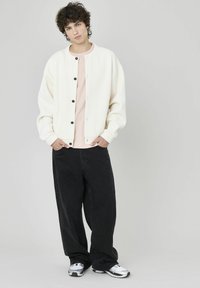 Veste bomber blanche texturée avec des boutons noirs, portée sur une chemise rose clair et accompagnée d'un pantalon noir ample et de baskets argentées.