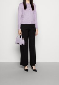 Cardigan lilas à boutons, associé à un pantalon large noir et des talons pointus noirs. Tenant un sac à main matelassé lavande.