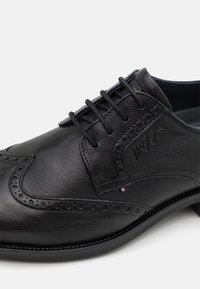 Chaussure en cuir noir avec détails brogues, bout rond et laçage. Surface texturée et subtile décoration de logo sur le côté.