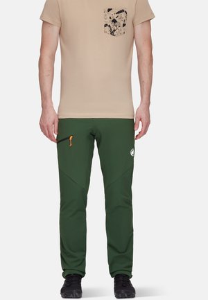Pantalons soft-shell verts avec des jambes fuselées, présentant une fermeture éclair orange, des accents noirs et un logo blanc. Associés à un t-shirt beige et une poche à motifs.