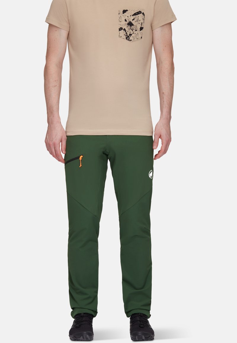 Groene zachte broek met taps toelopende benen, voorzien van een oranje rits, zwarte accenten en een wit logo. Gecombineerd met een beige t-shirt en een geprinte zak.