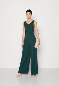 Combinaison verte sans manches avec un buste torsadé, design à jambes larges et texture lisse. Associée à une pochette argentée et des escarpins beiges à plateforme.