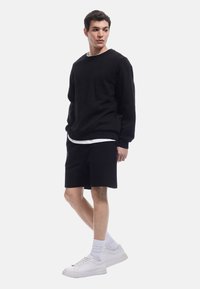 Sweatshirt en coton noir, shorts noirs ; chaussettes blanches mi-mollet et baskets. Le design présente une coupe décontractée et un look superposé.