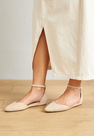 Ben som bär beige ankelremssandaler tillsammans med en krämfärgad midiklänning med en sidoklyfta, står på ett ljust trägolv.