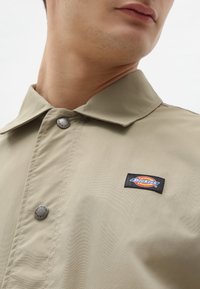 Beige jacka med krage, prydd med två metallknappar och en svart Dickies-logotyp på vänster sida. Slät textur, lättviktsmaterial.