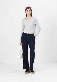 Lichtgrijs geribbeld langarmtop met een knoopsluiting, gecombineerd met marineblauwe wijde broek. Model houdt een witte handtas vast en draagt zwarte sneakers.