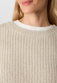 Beżowy sweter dziany z prążkowanym wzorem i subtelnymi srebrnymi akcentami, nałożony na białą koszulkę z okrągłym dekoltem. Zbliżenie na dekolt.