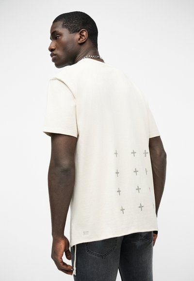 Άνδρας που φοράει εκρού μπλουζάκι με μικρούς γκρι σταυρούς τυπωμένους στο κάτω μέρος της πλάτης, συνδυασμένο με σκούρα γκρι τζιν και ασημένιο αλυσίδα κολιέ.