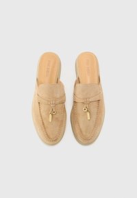 Beige suede slip-on shoes with a rounded toe, featuring a small decorative gold tag and a smooth, light-colored sole.