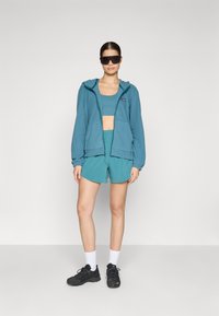 Teal hoodie with a front zipper, paired with a matching crop top and shorts. Textured fabric, worn with black shoes and large sunglasses.