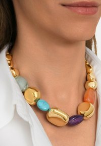 Collier avec de grandes pierres irrégulières en or, turquoise, violet, vert et orange porté avec une chemise blanche à col sur une femme.