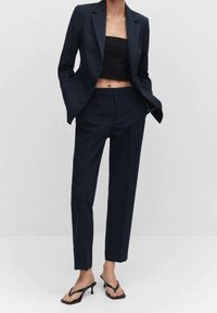 Costume bleu marine avec blazer ajusté et pantalon tailleur assorti, associé à un crop top noir et des tongs noires. Tissu lisse et design élégant.