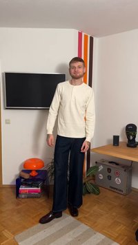 Ilgas rankovėmis kreminės spalvos henley marškinėliai, laisvo silueto tamsaus džinsinio audinio kelnės, poliruoti tamsūs batai, stovintis namų aplinkoje su spalvingais dekoratyviniais elementais.