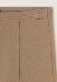 Leggings elasticizzati marroni con una texture liscia, caratterizzati da un piccolo logo in metallo e dettagli di cucitura visibili sulla vita.