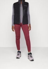 Black fleece vest with high collar, gray long-sleeve top, maroon leggings with stitching details, and gray and black sneakers.