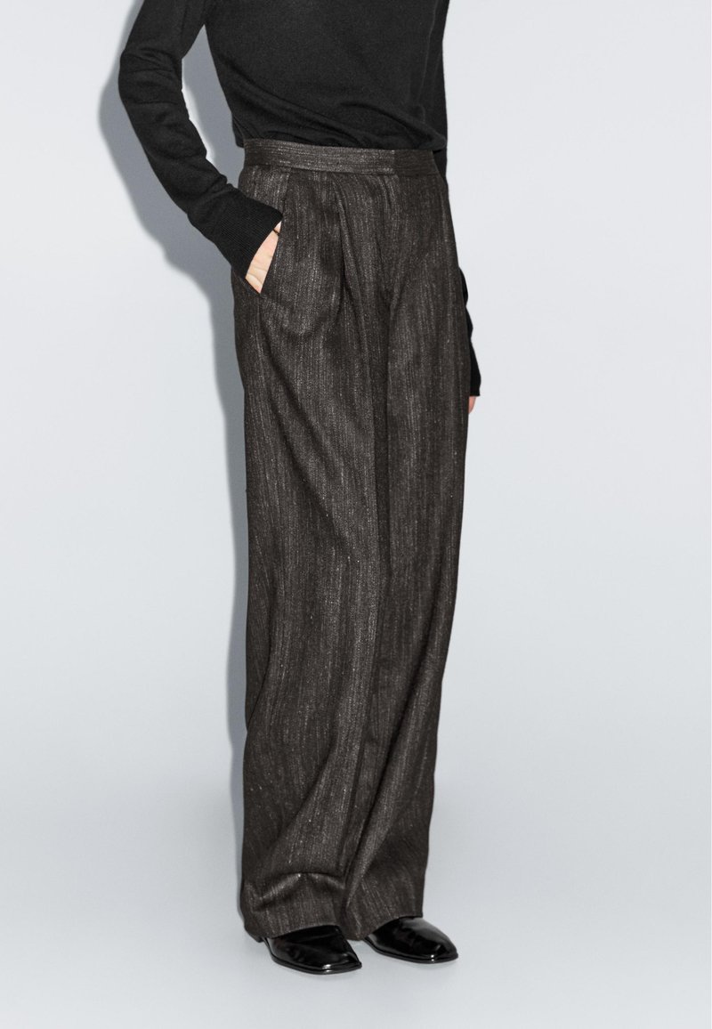 Wijde broek in donkergrijze textuurstof, voorzien van zijzakken, hoge taille en een geplooid ontwerp, gecombineerd met een zwarte longsleeve top.