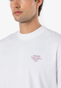 Camiseta de algodón blanca con cuello redondo. Presenta un pequeño logo rosa bordado en el lado izquierdo del pecho que dice "PEGADOR Certificado Auténtico AWG."