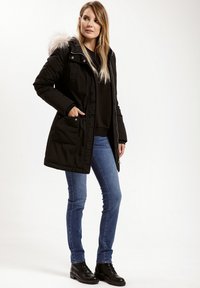 Czarna ocieplana parka z kapturem wykończonym sztucznym futrem, z zamkiem i napami, oraz kieszeniami z przodu, noszona na czarnym swetrze i niebieskich dżinsach.