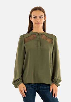 Blouse vert olive avec de longues manches bouffantes, accents en dentelle aux épaules, devant à boutons et tissu texturé. Portée avec un jean foncé.