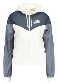 Nike huva jacka med dragkedja framtill, vit kropp och marinblå ärmar, tillverkad av lätt tyg med elastiska muddar.