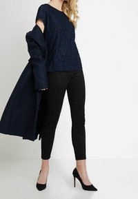 Granatowa koronkowa bluzka z krótkim rękawem, zestawiona z czarnymi zamszowymi spodniami. Modelka nosi czarne szpilki i trzyma granatowy płaszcz na ramieniu.