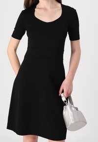 Černé šaty s krátkými rukávy, kulatým výstřihem, upnutým pasem a rozšířeným lemem. Doplněné malou stříbrnou kabelkou. Hladká textura.