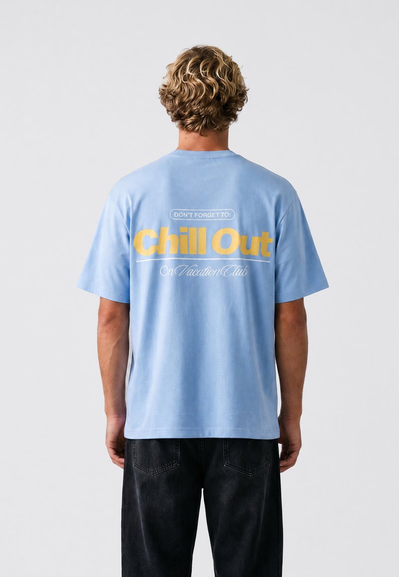 Personne blonde aux cheveux courts et bouclés porte un t-shirt bleu clair avec un texte jaune « Chill Out » vu de dos sur un fond uni.