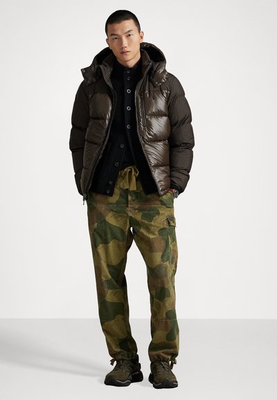 Brown puffer jacket with a hood, layered over a black button-up shirt. Olive camouflage cargo pants with drawstring cuffs. Dark athletic shoes.