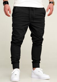 Black jogger pants with an elastic waistband and cuffed ankles. Made of soft material, featuring side pockets and minimal seams. Worn with white sneakers.
