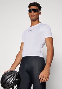 Maillot en maille à manches courtes de couleur blanche, associé à un cuissard de cyclisme noir. Le modèle tient un casque noir ; les caractéristiques incluent un design ajusté et des détails de logo.