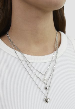 Neck with four layered silver chains featuring a pearl, a sun pendant, and a heart pendant over a white ribbed top.