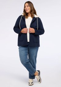 Giacca blu marina con cappuccio crema e lacci, dotata di due tasche frontali e una vestibilità comoda, abbinata a jeans azzurri chiari.