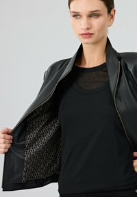 Giubbotto di pelle nera aperto per rivelare un interno a motivo scuro; modello che indossa un top nero a strati. Texture liscia, design elegante.