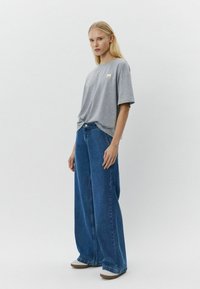 Camiseta de algodón gris con manga corta y un pequeño parche con logo; combinada con unos vaqueros de pierna ancha de denim azul y zapatos de lona beige sin cordones.