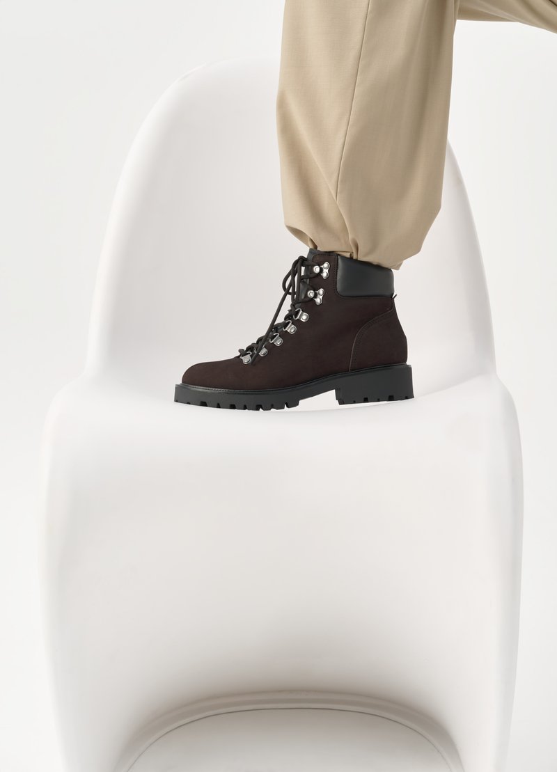 Bruine suède enkellaars met zwarte leren accenten, ronde neus, vetersluiting aan de voorkant met metalen hardware, en robuuste zwarte zool, tentoongesteld op een stoel.