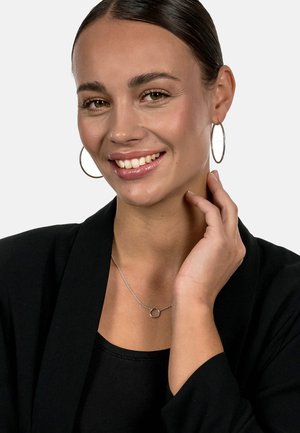 Silberne Creolen und eine zarte Halskette mit einem runden Anhänger. Modell trägt ein schwarzes Oberteil und eine Jacke, lächelt mit der Hand am Hals.