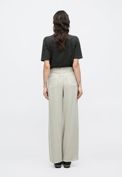 Γκρι κοντομάνικο πουκάμισο συνδυασμένο με μπεζ παντελόνια ψηλόμεσα, φαρδιά, με πίσω τσέπες και λεία υφή. Το σύνολο ολοκληρώνουν μαύρα παπούτσια.