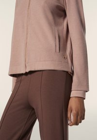 Giacca rosa chiaro con finitura testurizzata, dotata di cerniera e tasche laterali, abbinata a pantaloni marroni aderenti. Dettaglio in hardware bronzo visibile.