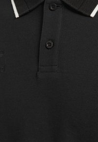 Svart polo skjorta med texturerad tyg, tre-knapp placket och en kontrasterande vit rand på kragen. Diskret logotypdetalj finns.