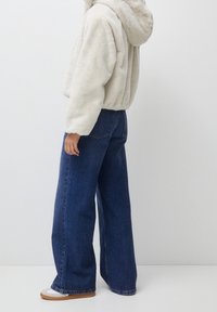 Veste à capuche en fausse fourrure couleur crème, associée à un jean large taille haute bleu foncé. Les chaussures présentent un schéma de couleurs blanc et fauve.